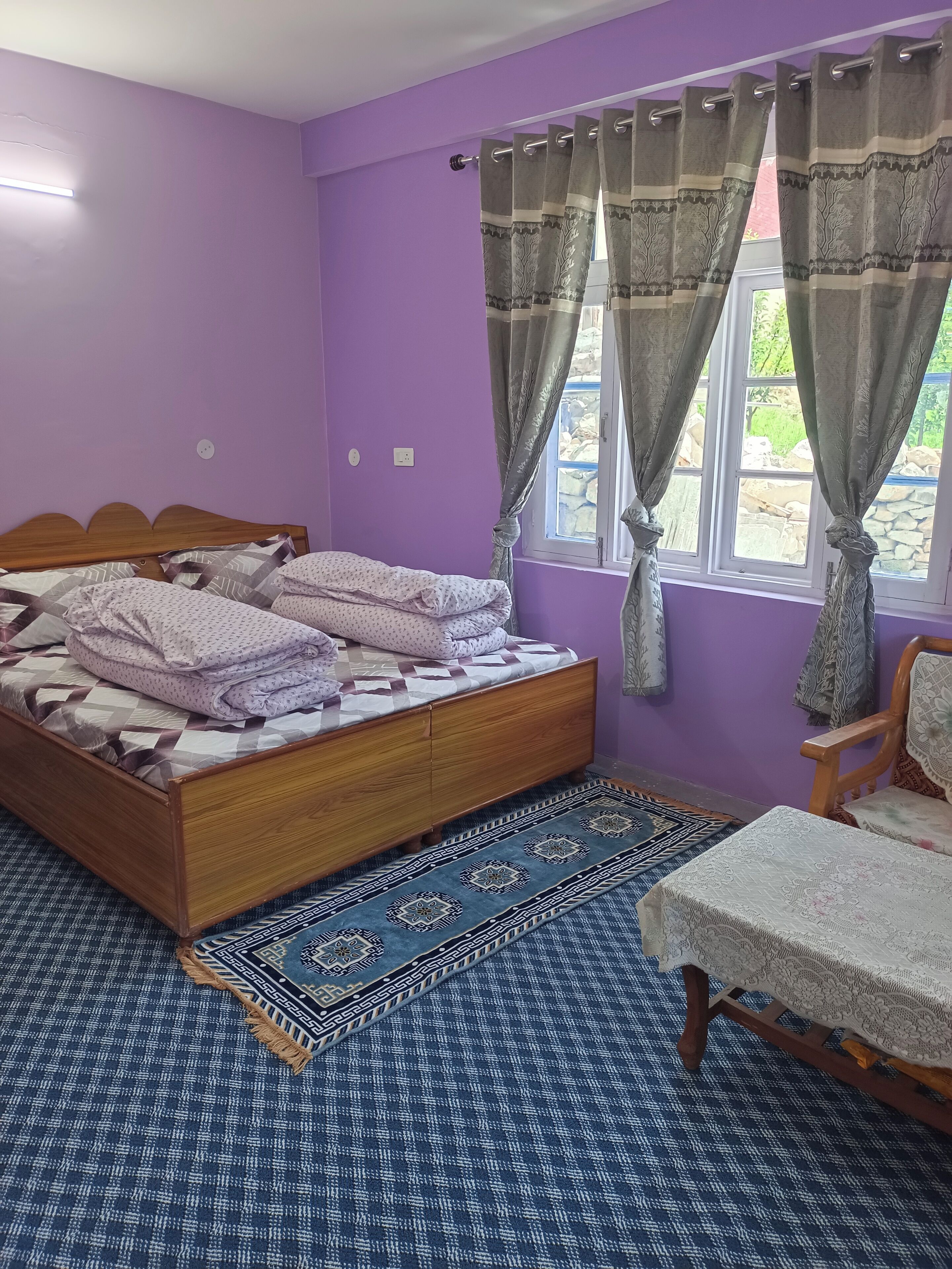 A bedroom with a wooden bed, purple walls, a window with curtains, and a patterned rug.
