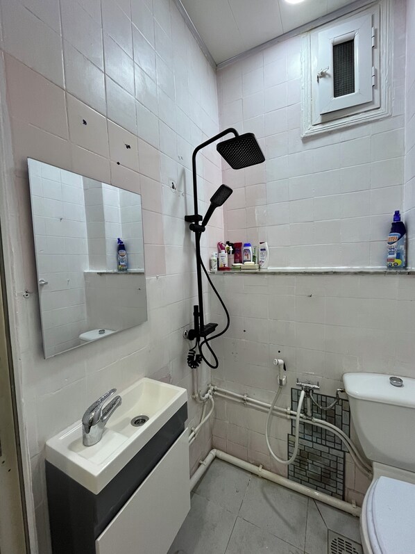 A bathroom with a white sink, mirror, and toilet. There is a showerhead mounted on the wall, and toiletries are visible on a shelf. - Cosy F2 apartment in the heart of Algiers (Alger Centre)
