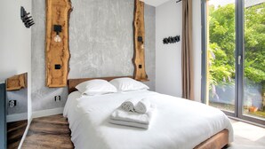 Une chambre moderne avec un grand lit, une tête de lit en bois et une vue sur la verdure par la fenêtre.