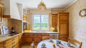 Een keuken met houten kastjes, een eettafel met stoelen, een kroonluchter en een raam met uitzicht op groen.