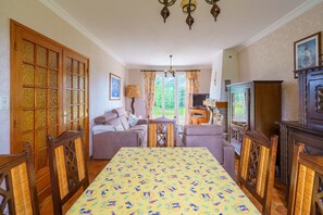 Ein Essbereich mit einem Tisch, gedeckt für vier Personen, umgeben von Holzstühlen mit gewobenen Rückenlehnen. Eine Kristallleuchter hängt von der Decke, und ein Fernseher ist an der Wand angebracht.