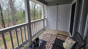 A covered porch with a table and chairs, a door, and a view of the surrounding forest. - Lake Escape Getaway (Fairfield Bay)