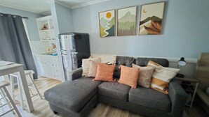 A living room with a grey sectional sofa, a white table, and a kitchen area with a refrigerator. - Lake Escape Getaway (Fairfield Bay)