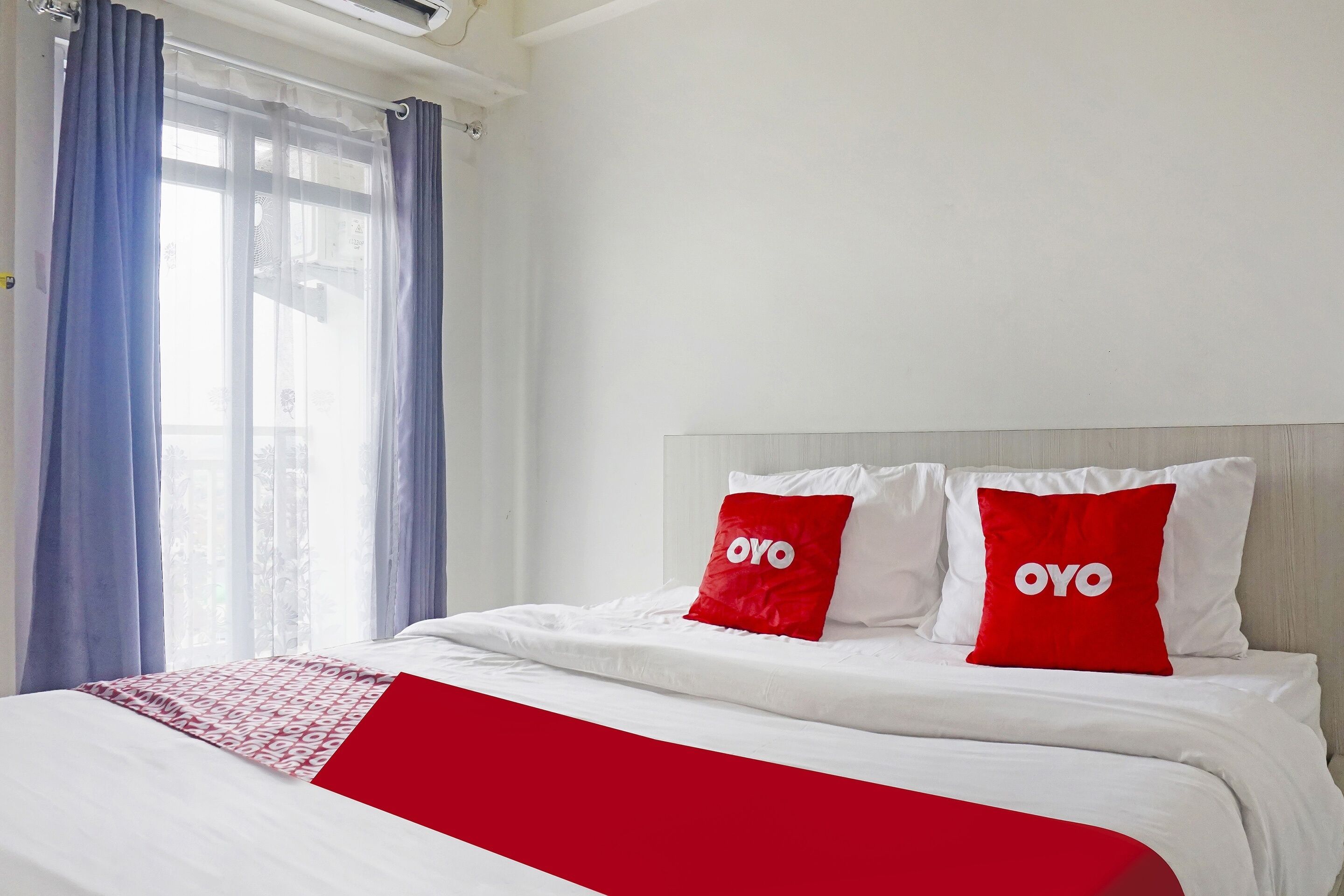 Une chambre d’hôtel avec un lit, du linge de lit blanc, des oreillers rouges ornés du mot « OYO » et une fenêtre avec des rideaux bleus.