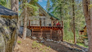 Eine Holzhütte mit einer Holzterrasse, großen Fenstern und einem geneigten Dach, umgeben von Bäumen und Felsen.