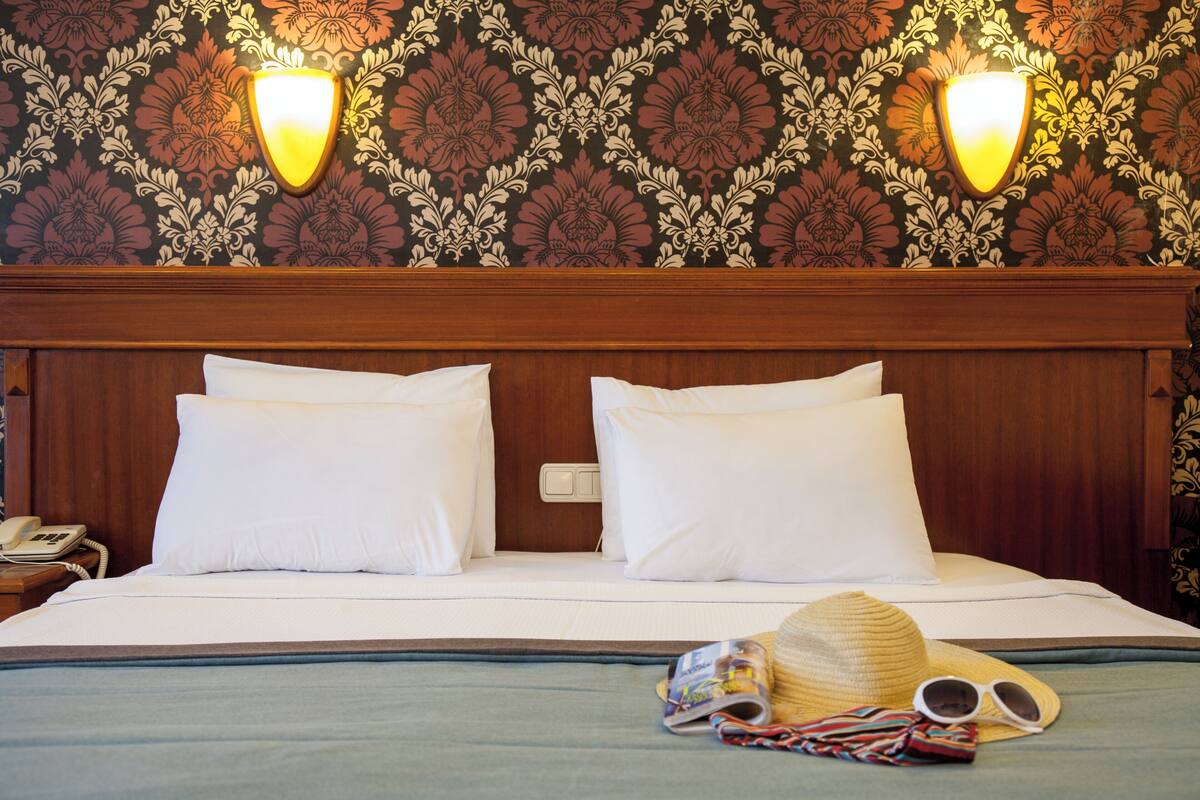 A hotel room with a wooden headboard, two white pillows, a bed with a blue bedspread, a telephone on the nightstand, and a hat and sunglasses on the bed.