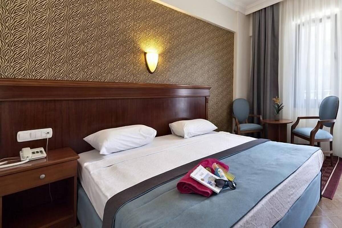 A hotel room with a large bed, a bedside table with a phone, a wall-mounted light, a small table with chairs, and a view of the outdoors through sheer curtains.