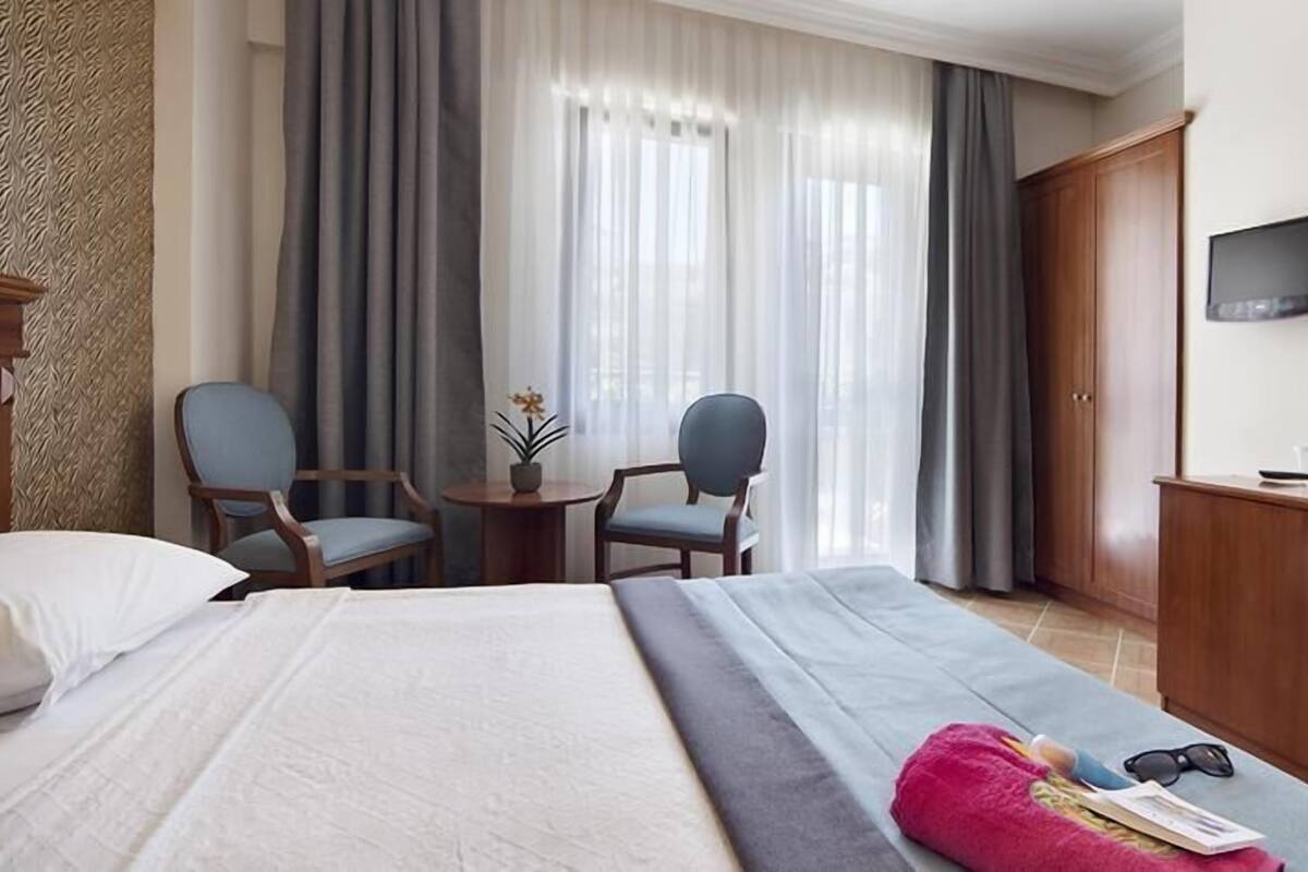 A hotel room with a bed, two chairs, a desk, a TV, and a small table with a plant.