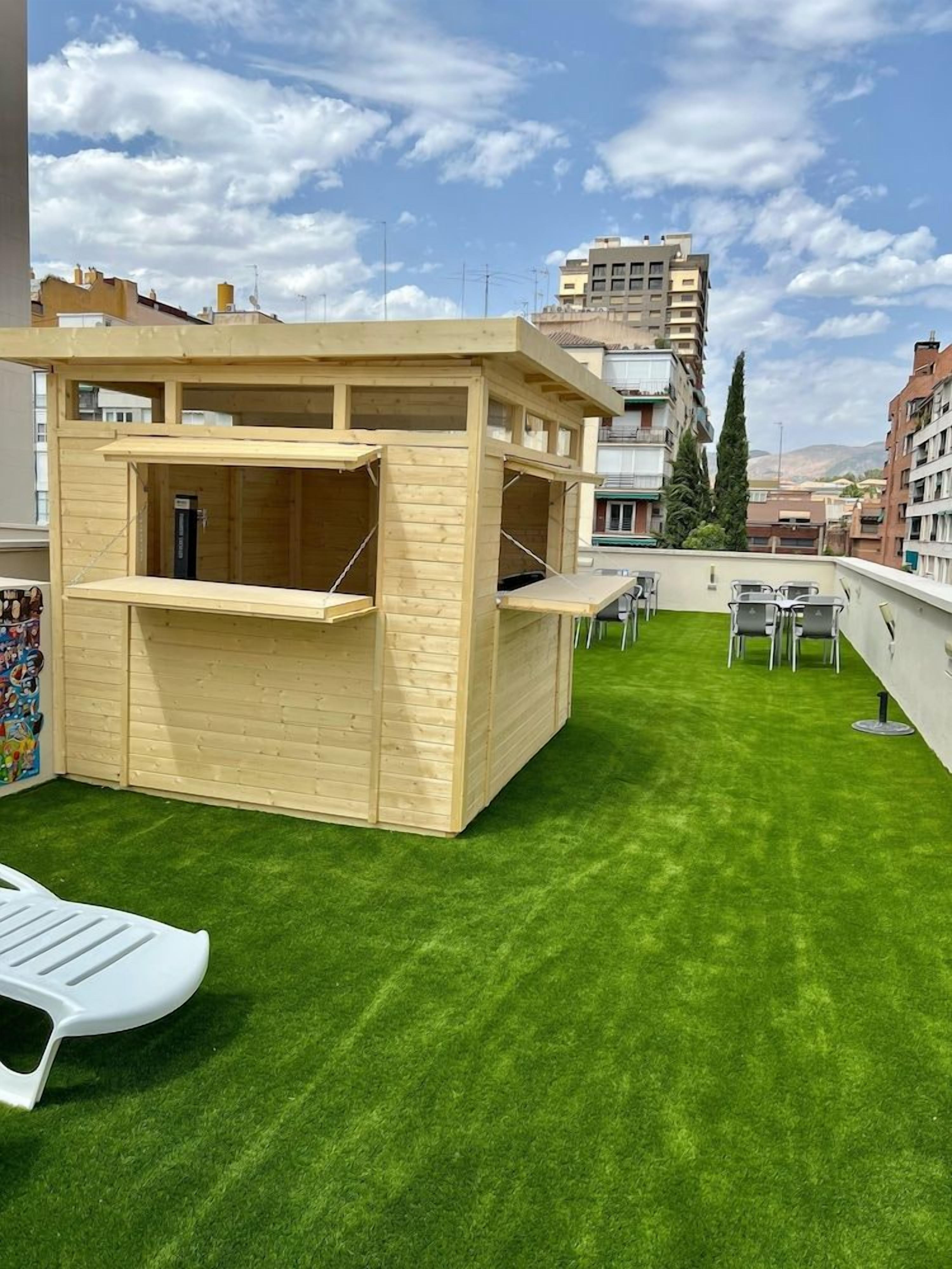 A rooftop with artificial grass, a wooden play structure, and a seating area with tables and chairs.