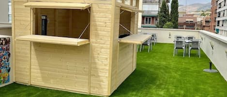 A rooftop with artificial grass, a wooden play structure, and a seating area with tables and chairs.
