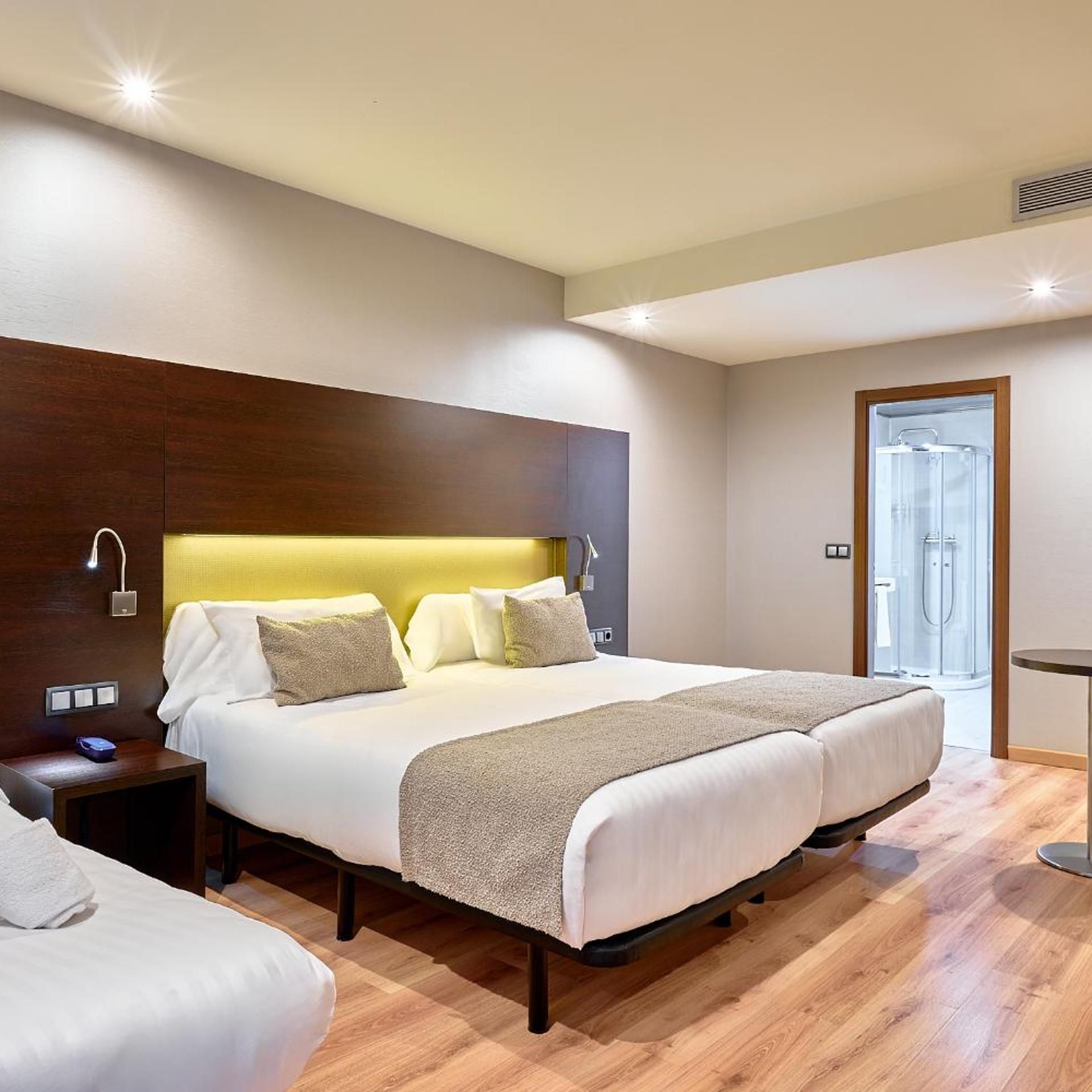 Une chambre d’hôtel moderne avec un grand lit, des tables de chevet, une tête de lit en bois et une porte de douche en verre.