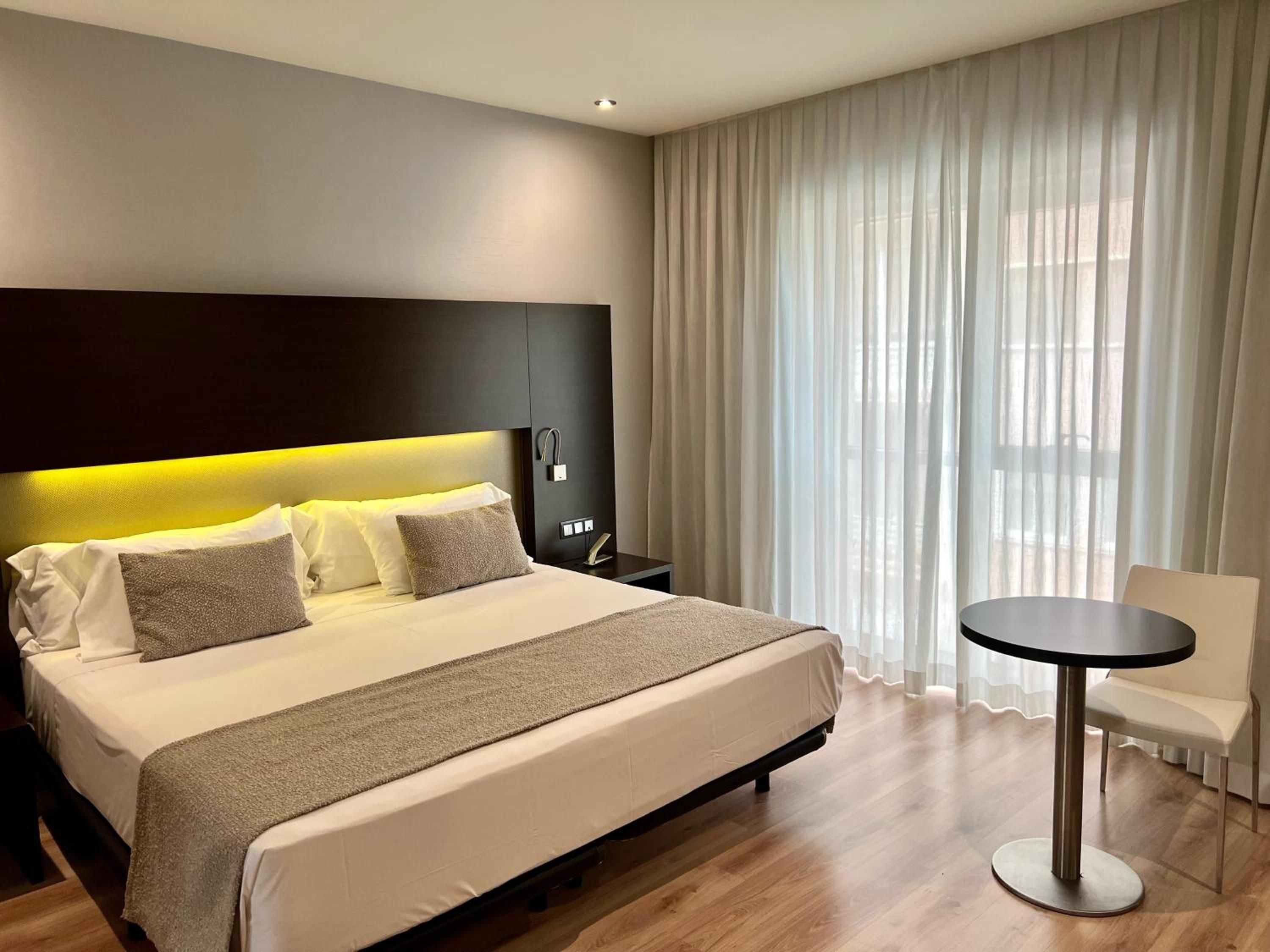 A modern hotel room with a large bed, a bedside table, a chair, and a window with sheer curtains.