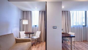 Una habitación de hotel moderna con un sofá, una mesa de comedor con sillas, una lámpara y ventanas amplias con cortinas.