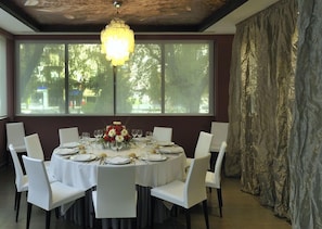 A round dining table set for six with white chairs, a chandelier, and a view of trees through large windows. - Hotel Turia Granada (Granada)