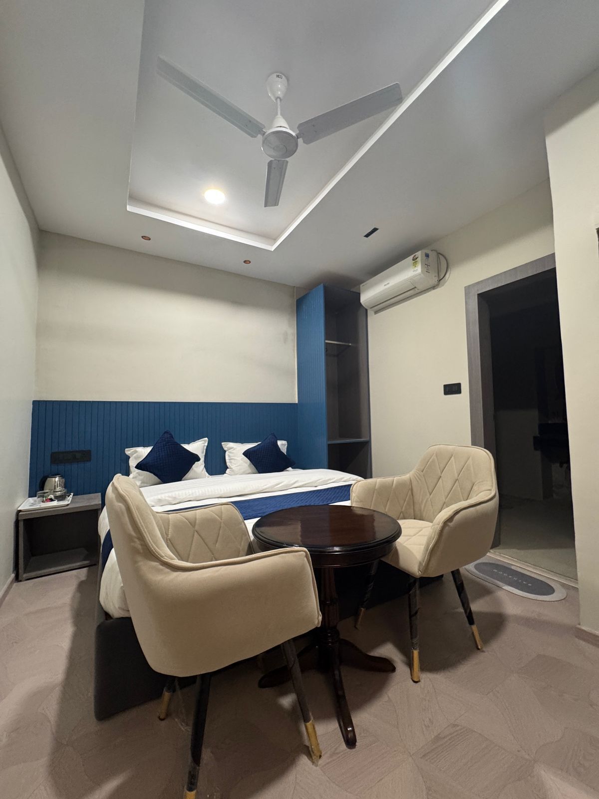 A modern hotel room with a bed, two chairs, a small table, a ceiling fan, and an air conditioning unit.