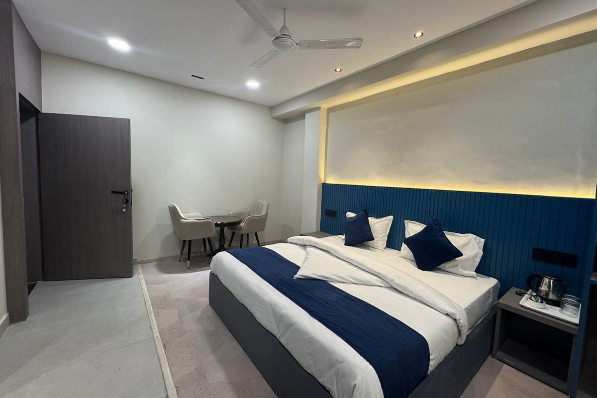 A modern hotel room with a large bed, a desk with a chair, a ceiling fan, and a nightstand with a coffee pot.