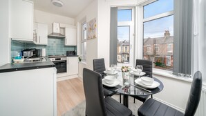 Una cocina moderna con armarios blancos, un comedor con mesa de vidrio y vistas a la ciudad a través de grandes ventanales.