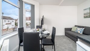 Una sala de estar moderna con una mesa de comedor lista para cuatro personas, un sofá gris y un gran ventanal con vistas a la calle.