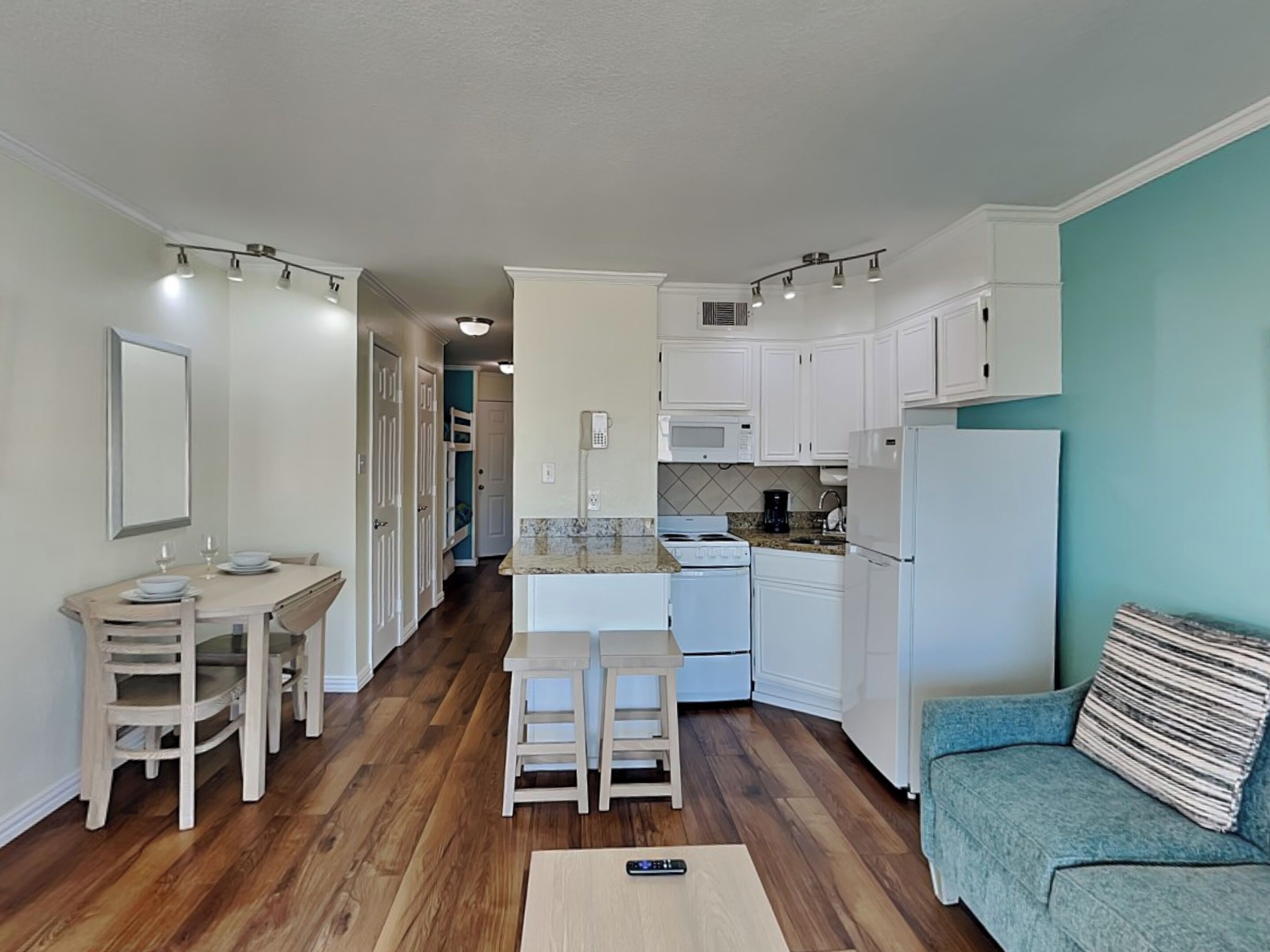 A compact kitchen with white cabinets, a dining area with wooden chairs, and a teal-colored sofa.
