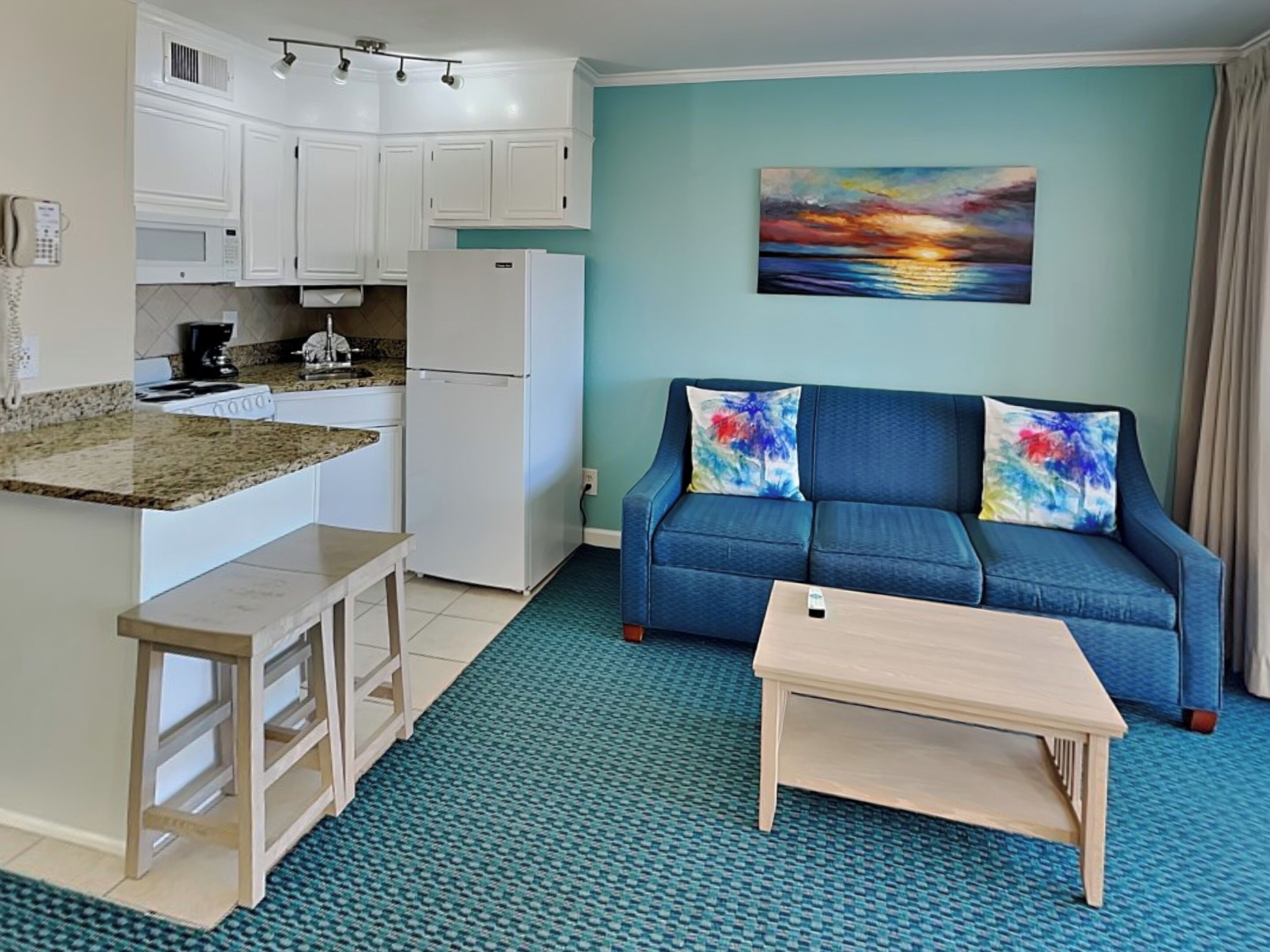 Un espacio habitable compacto con kitchenette, un sofá azul con cojines de colores y una pintura de un atardecer.