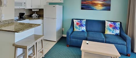 Un espacio habitable compacto con kitchenette, un sofá azul con cojines de colores y una pintura de un atardecer.