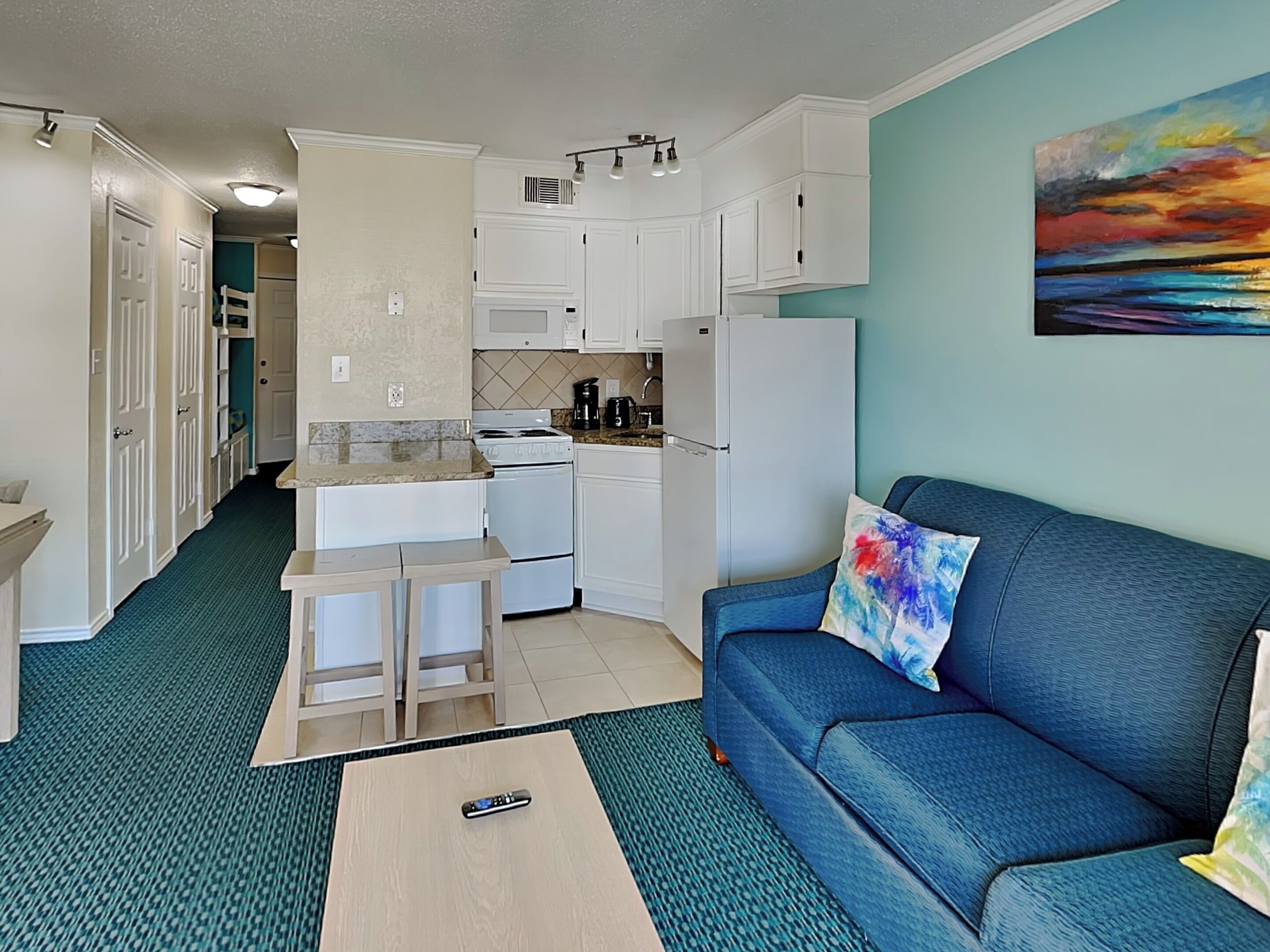 A compact kitchen with white cabinets, a microwave, stove, and refrigerator. There's a blue sofa with a colorful pillow and a remote control on a small table.