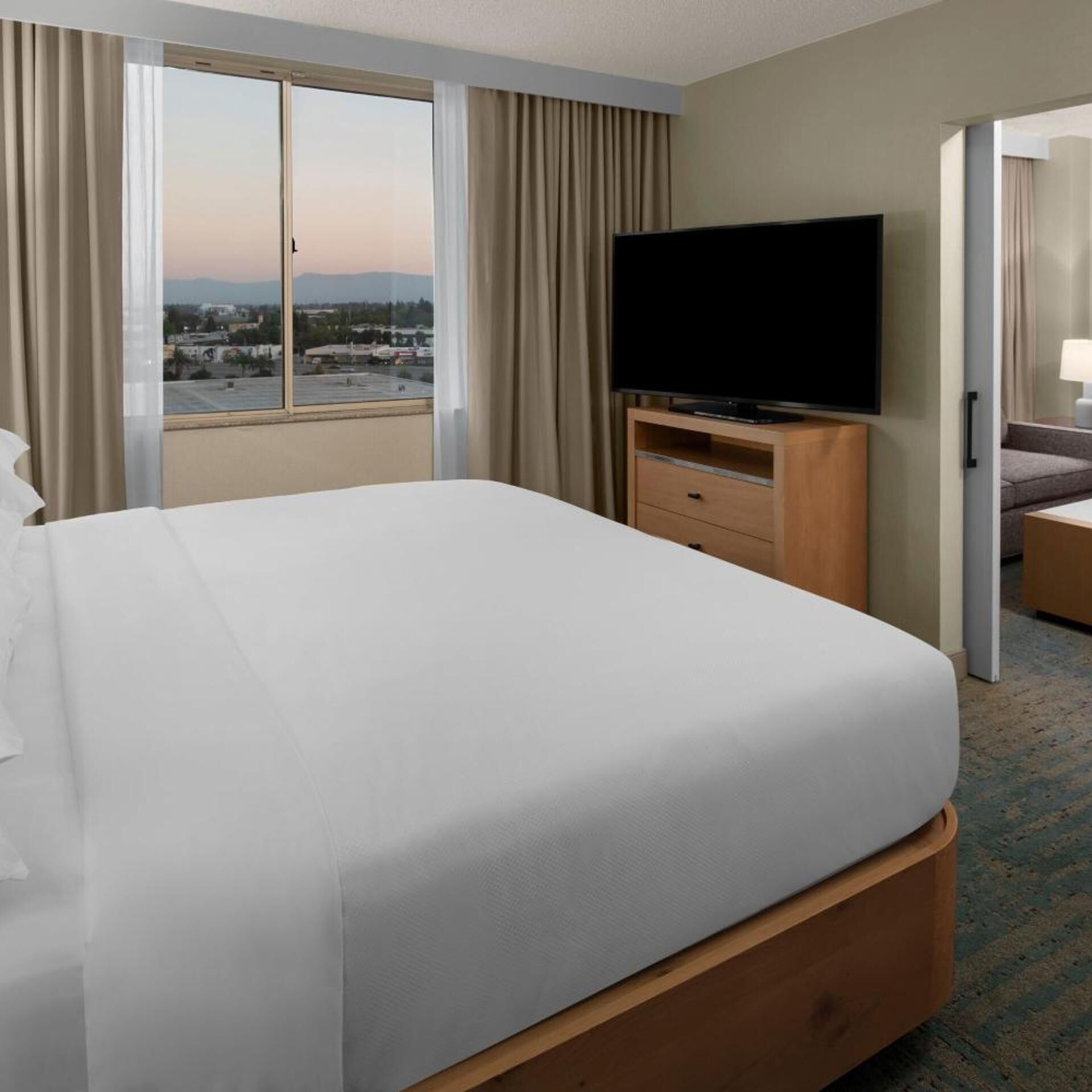 A hotel room with a large bed, a television on a wooden stand, and a view of a cityscape through the window.