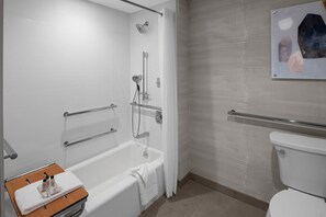 Egy modern fürőszoba, amelyben kád, zuhanyzó és WC található. Emelőkorlátok és törölközőtartó is van.