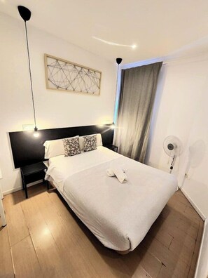 A modern hotel room with a large bed, a black headboard, a bedside table, a floor lamp, a ceiling light, a window with curtains, and a wall-mounted fan. - A&Z Javier Cabrini (Madrid)