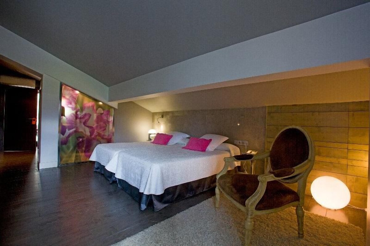 A bedroom with a bed, a chair, a wall with a light, and a door.