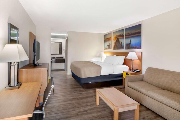 A hotel room with a bed, a sofa, a desk, a TV, and a painting of a sunset. - Quality Inn & Suites Decatur - Priceville I-65 (Decatur)