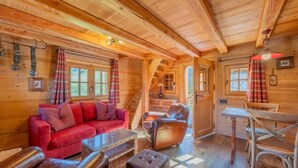 Interior acogedor de una cabaña de troncos con un sofá rojo, sillones de cuero, una mesa de comedor de madera y una escalera que conduce a un nivel superior.