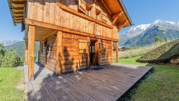 Una cabaña de madera con terraza, puerta principal y vista a la montaña.