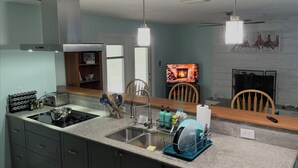 Une cuisine équipée d’une cuisinière, d’un évier et d’un égouttoir. On y trouve une table à manger en bois avec des chaises. Une cheminée est visible au fond de la pièce.