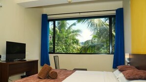 A hotel room with a bed, a desk with a television, and a window with curtains. - Relax Holiday Home (Madgaon)