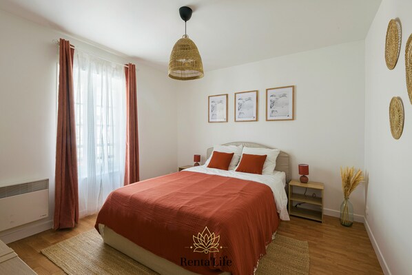 Un dormitorio con cama, mesita de noche, lámpara, jarrón con plantas secas y cuadros en la pared.