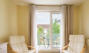Une pièce comprenant deux chaises blanches, une petite table et une porte-fenêtre coulissante donnant sur un balcon extérieur équipé de chaises.