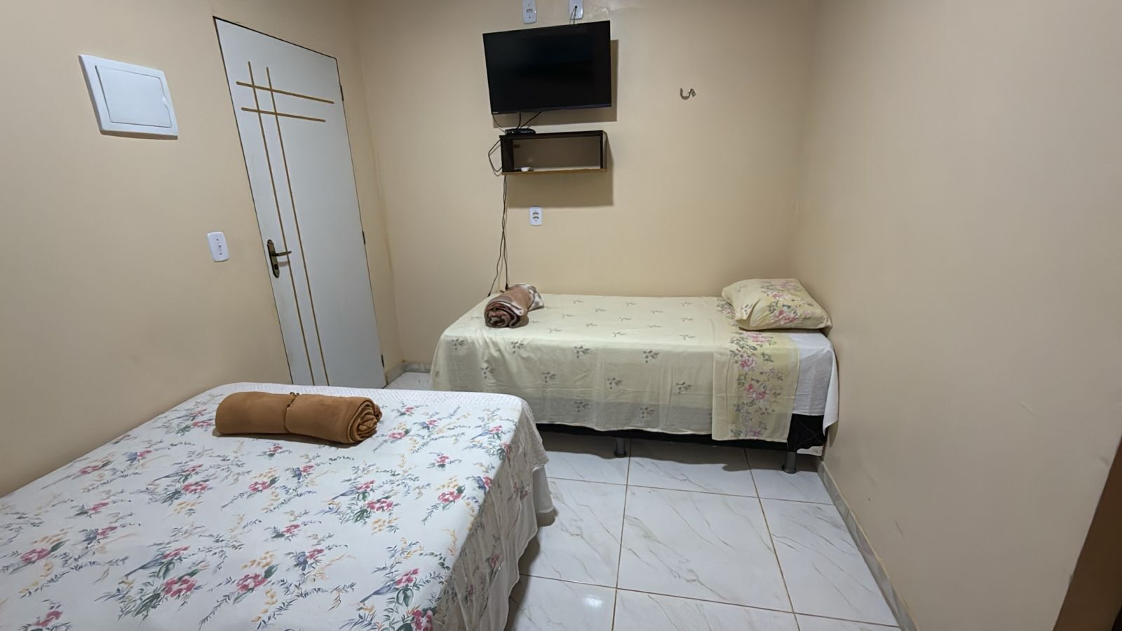 Een kleine, schone kamer met twee eenpersoonsbedden, een aan de muur bevestigde televisie en een deur met een hendel.