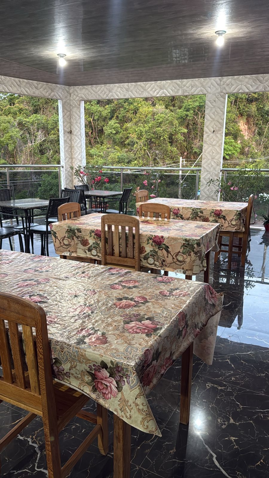 An outdoor dining area with tables covered in floral tablecloths, wooden chairs, and a glass railing.
