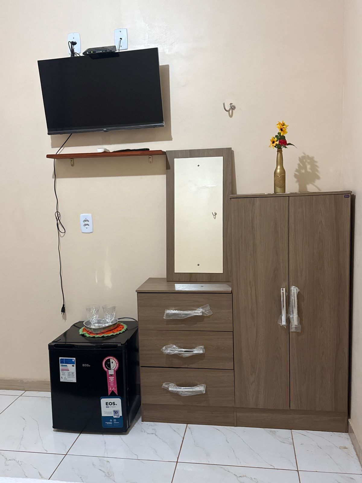 A room with a TV mounted on the wall, a wooden cabinet with drawers, a small table with a vase of flowers, and a black box with a sticker.
