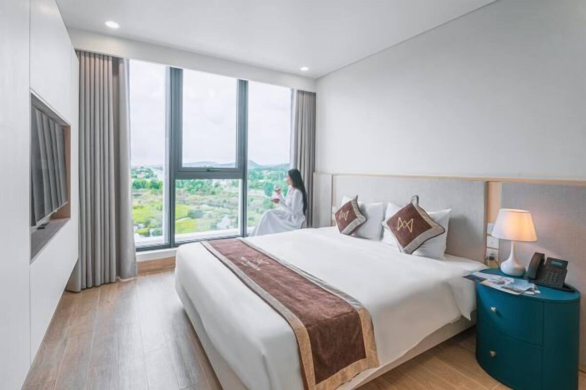 A hotel room with a large bed, a view of the city, a desk with a lamp, and a TV mounted on the wall.