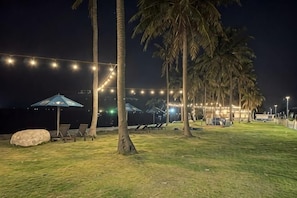 Un espacio al aire libre de noche con luces colgantes, palmeras y sillones de descanso.