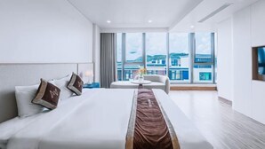 A modern hotel room with a large bed, a view of the city through large windows, and a small seating area with a table. - Mey Mansion Hotel Phu Quoc (Phu Quoc Island)