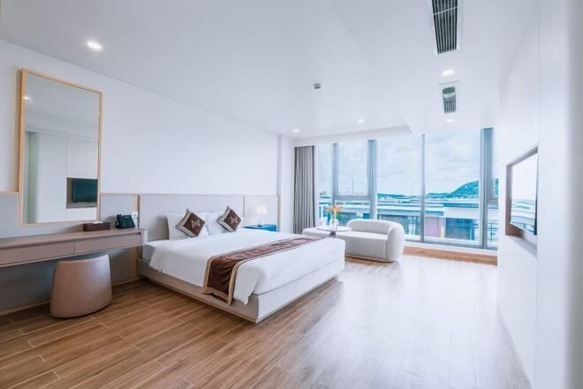 A modern hotel room with a large bed, a desk, a chair, and a large window offering a city view.
