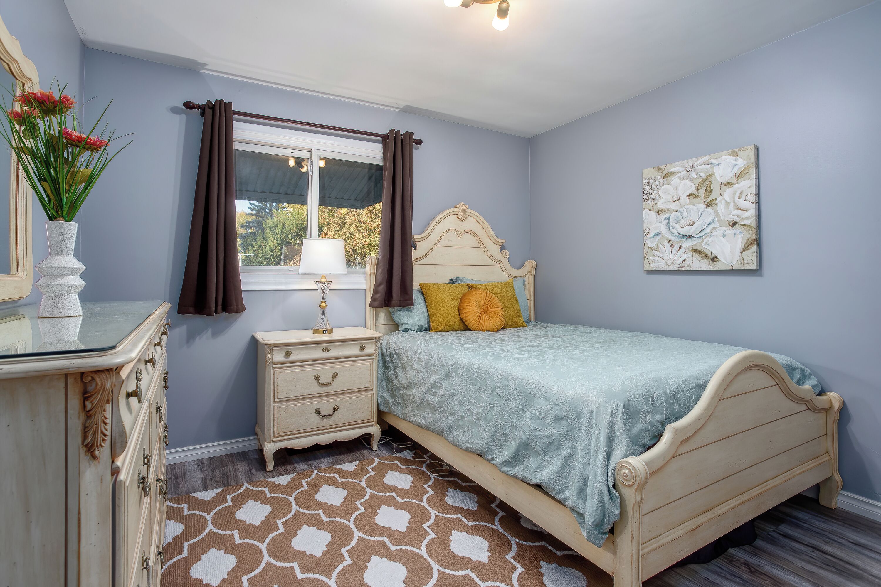 Un dormitorio con cama, cómoda, mesita de noche, lámpara y ventana con cortinas.