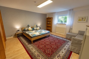 Une chambre à coucher avec un lit en bois, un tapis rouge à motifs, une bibliothèque et une fenêtre avec des rideaux.