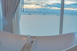 Une salle de bain moderne avec une baignoire sur pieds, une fenêtre donnant sur la mer et une petite étagère où sont rangés les articles de toilette.