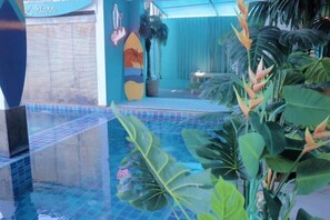 Un espace piscine agrémenté d’une décoration sur le thème du surf, de plantes tropicales et d’un sol carrelé.