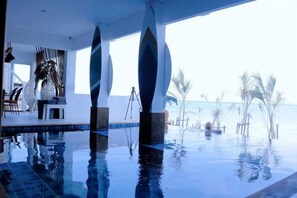 Piscina infinita en interior con vistas despejadas al océano, rodeada de sillones de descanso y palmeras.