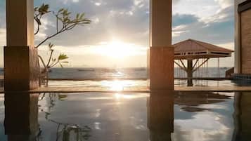 Una piscina infinita que refleja el sol y la vista al océano, con una estructura de madera y una planta en primer plano.